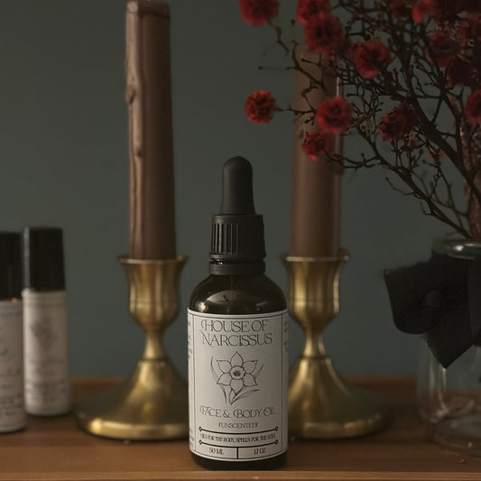 Bottle of House of Narcissus face and body oil on a wooden surface with candles and flowers in the background.