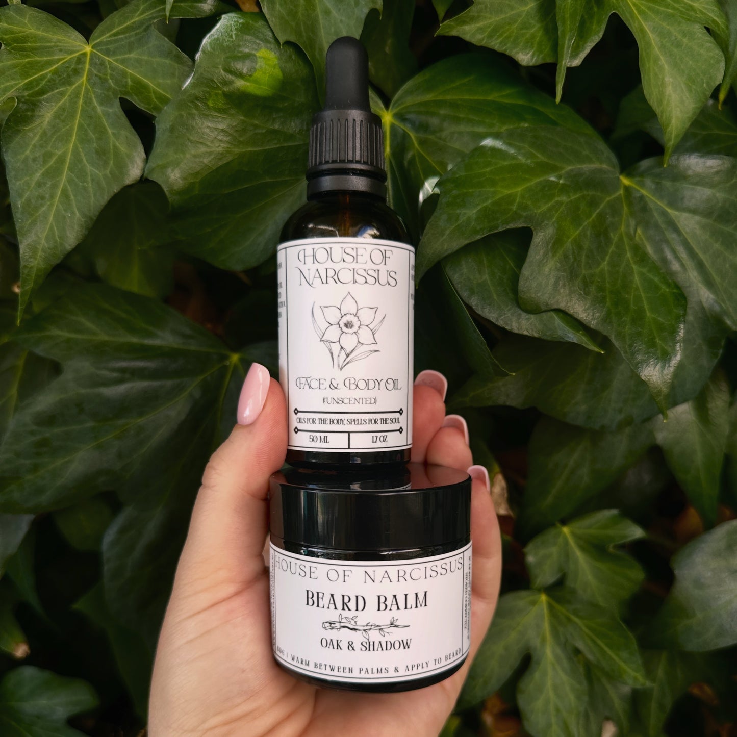 Hand holding two bottles of 'House of Narcissus' products against a green leafy background. The Face & Body Oil, and the Beard Balm products.
