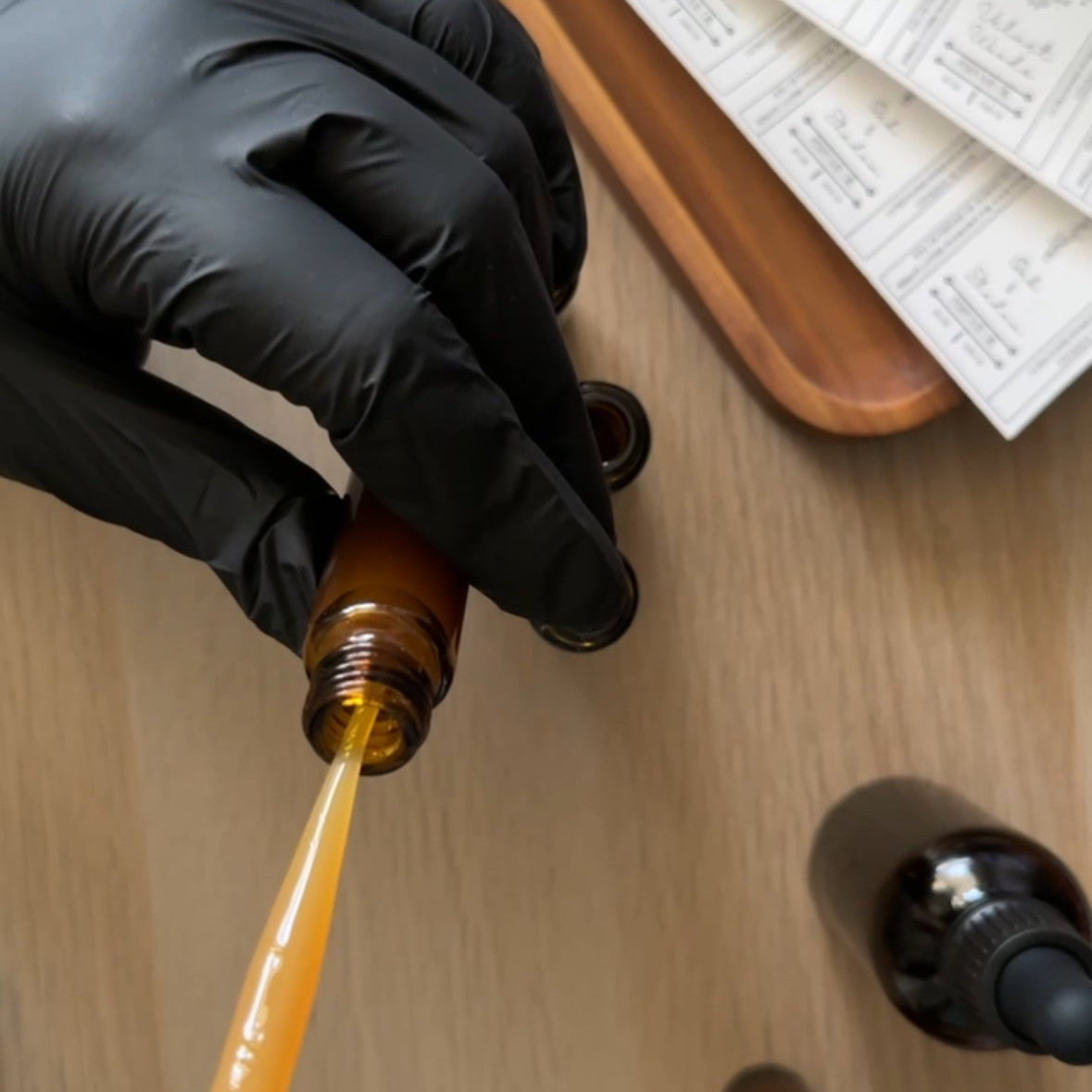 Person in black gloves pouring liquid from a brown bottle into another container on a wooden surface.