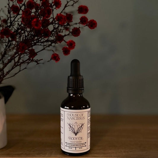Bottle of 'House of Narcissus' body oil on a wooden surface with red flowers in the background.
